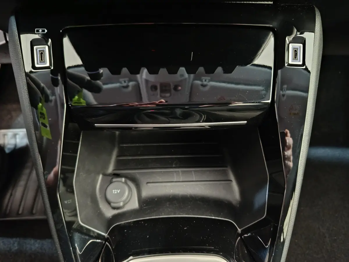Gros plan sur la console centrale noire brillante de la Peugeot 208 PureTech 100 S&S Allure 2025 avec prise 12V et ports USB.