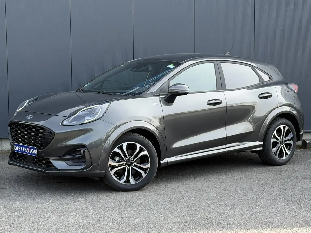 Ford Puma Ecoboost 155 ST Line gris magnétique en 3/4 avant droit, avec jantes alliage et calandre noire distinctive.