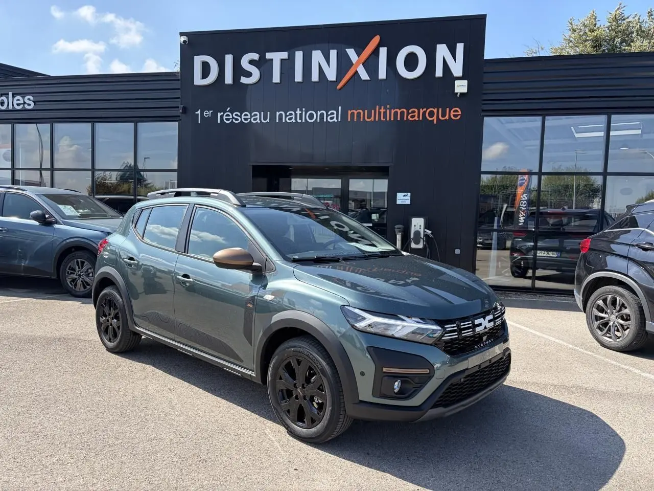 Vue 3/4 avant droite d'une Dacia Sandero III Stepway Vert Cèdre avec jantes noires et rétroviseurs cuivrés, devant un showroom Distinxion.