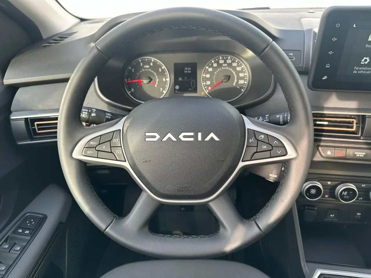 Vue rapprochée du volant noir Dacia avec commandes intégrées et tableau de bord numérique en arrière-plan.