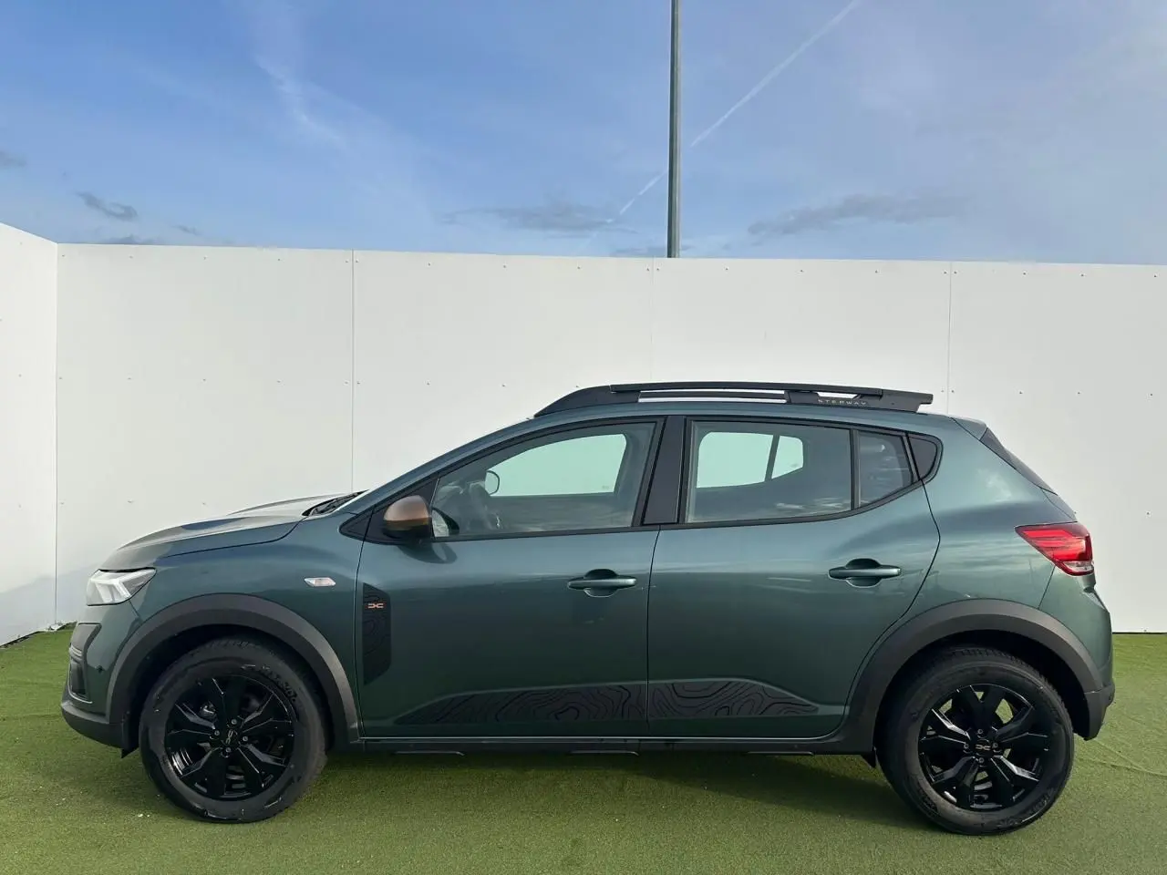 Profil latéral gauche de la Dacia Sandero Stepway 2025 en vert cèdre avec jantes noires et barres de toit brun cuivré.