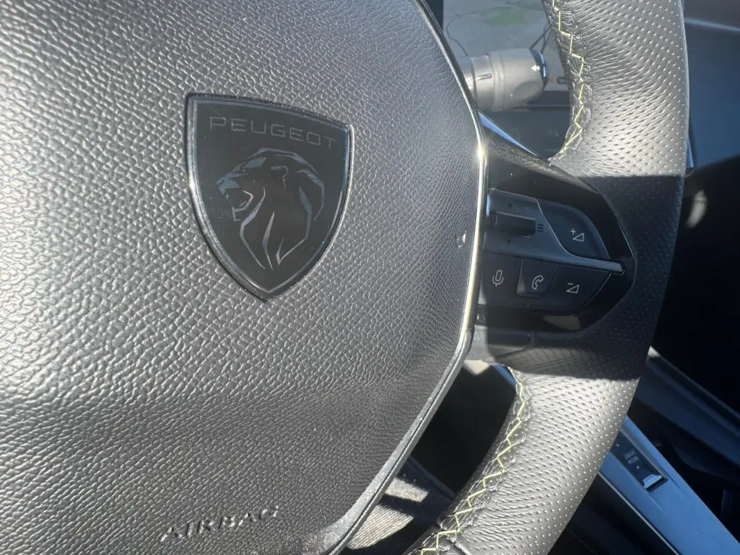 Gros plan sur le volant cuir noir perforé de la Peugeot 408 Hybrid 136 e-DCS6 GT avec logo lion et commandes intégrées.