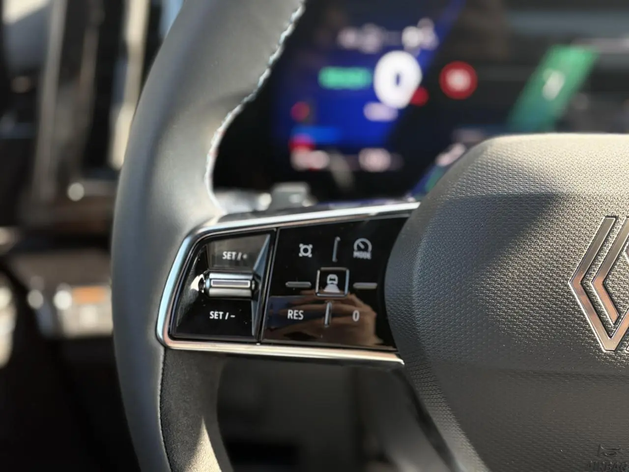 Gros plan sur les commandes gauche du volant gris texturé du Renault Austral E-Tech, avec tableau de bord numérique flou en arrière-plan.