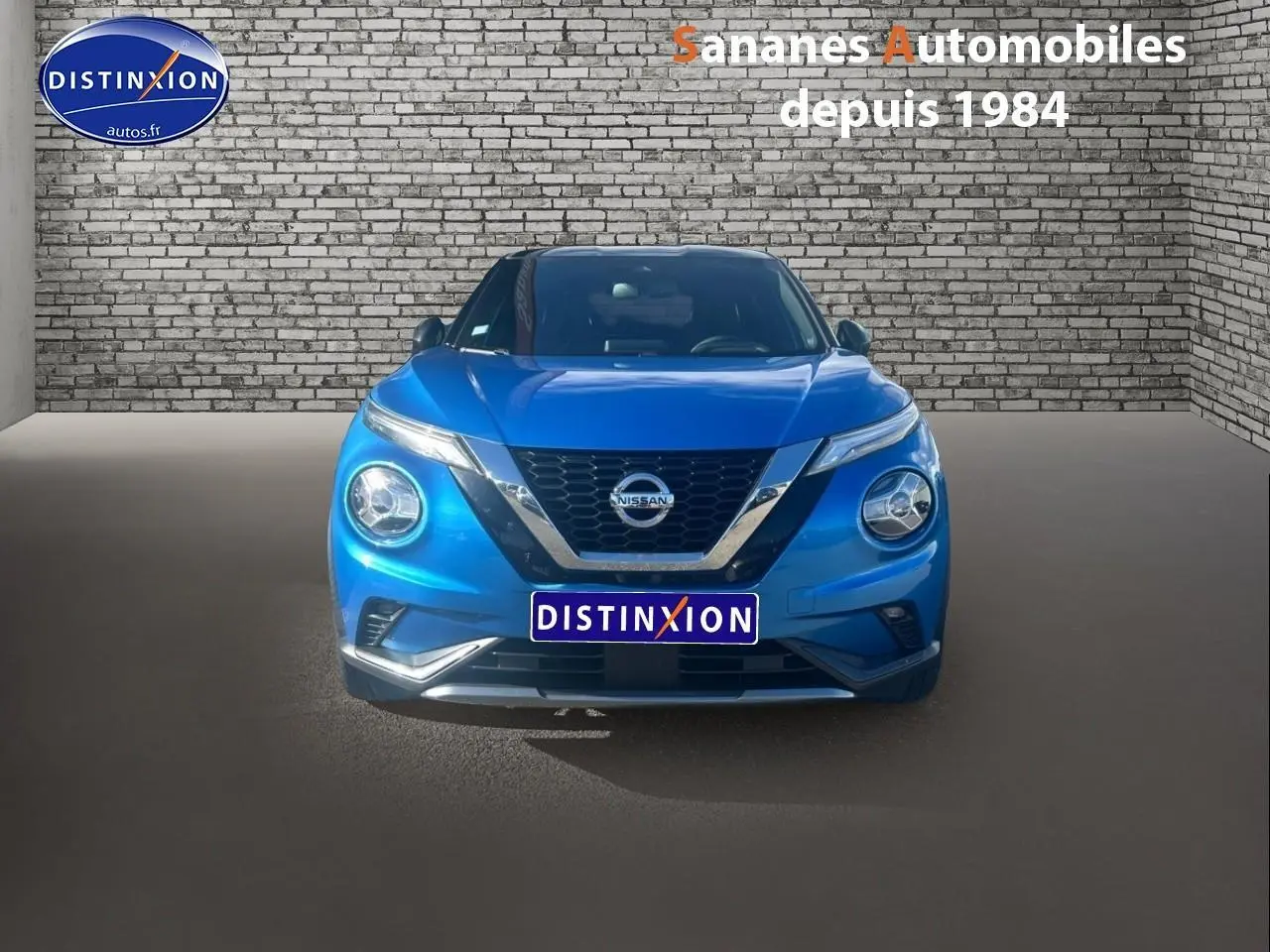 Vue avant d'un Nissan Juke 2020 bleu clair avec calandre chromée et phares allumés sur fond de mur en briques.