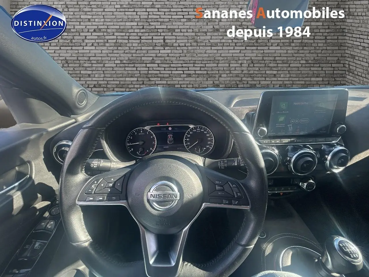 Vue intérieure centrée sur le volant Nissan d’un Juke bleu clair, avec tableau de bord et écran tactile visible.
