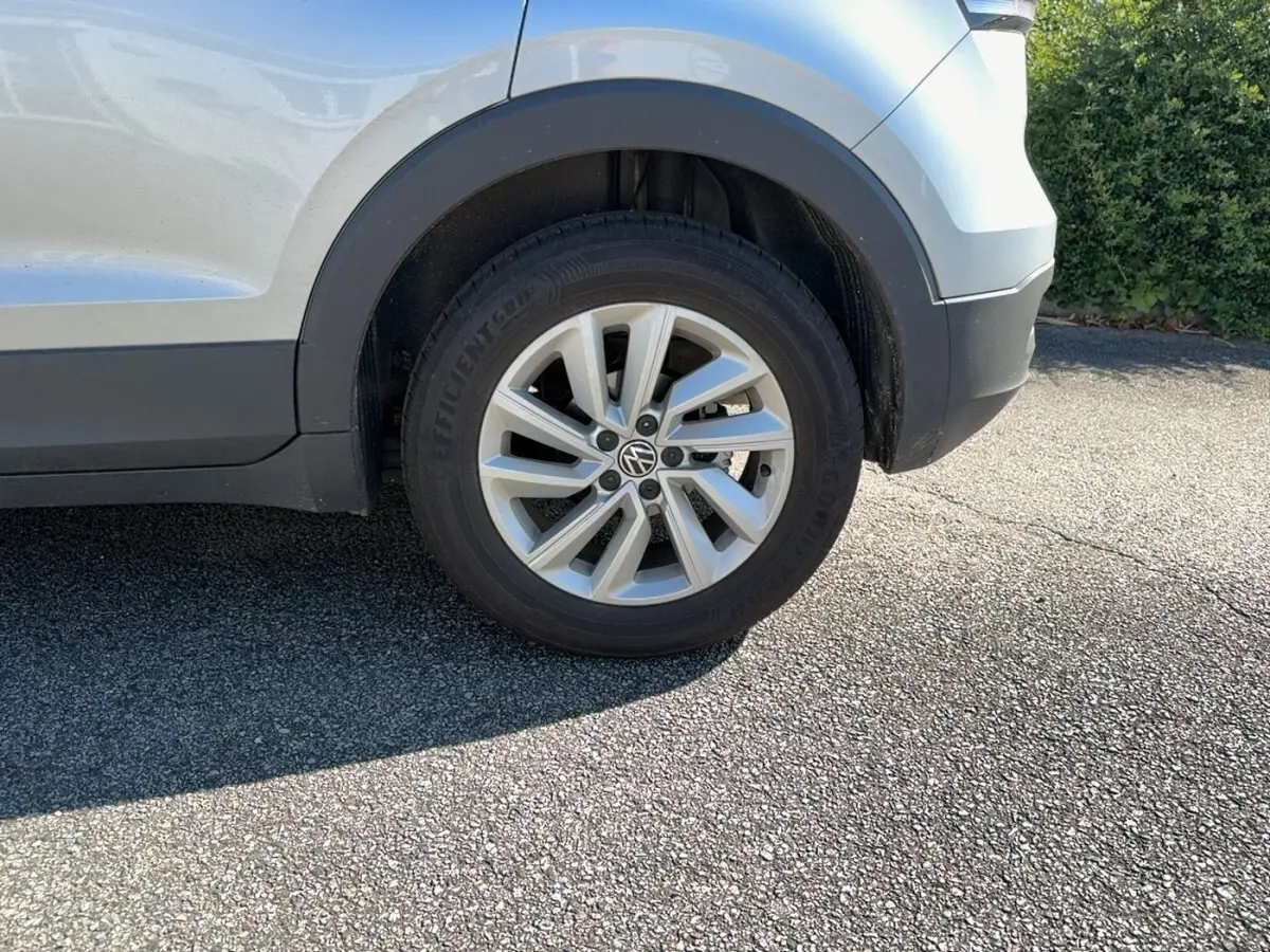 Gros plan sur la roue avant droite d’un Volkswagen T-Cross gris clair avec jante alliage et logo VW visible.