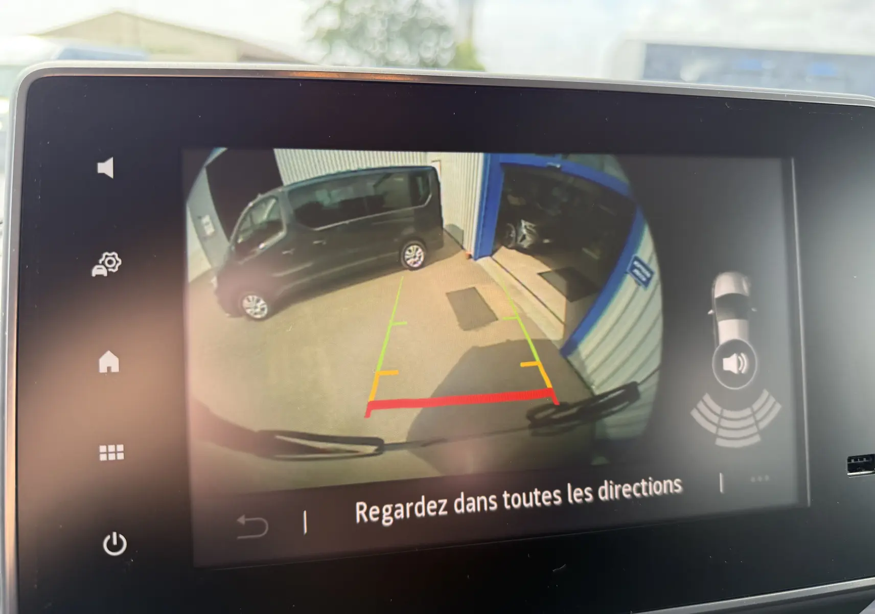 Vue caméra de recul montrant un Renault Trafic Combi gris Urban garé côté gauche près d'un bâtiment bleu.