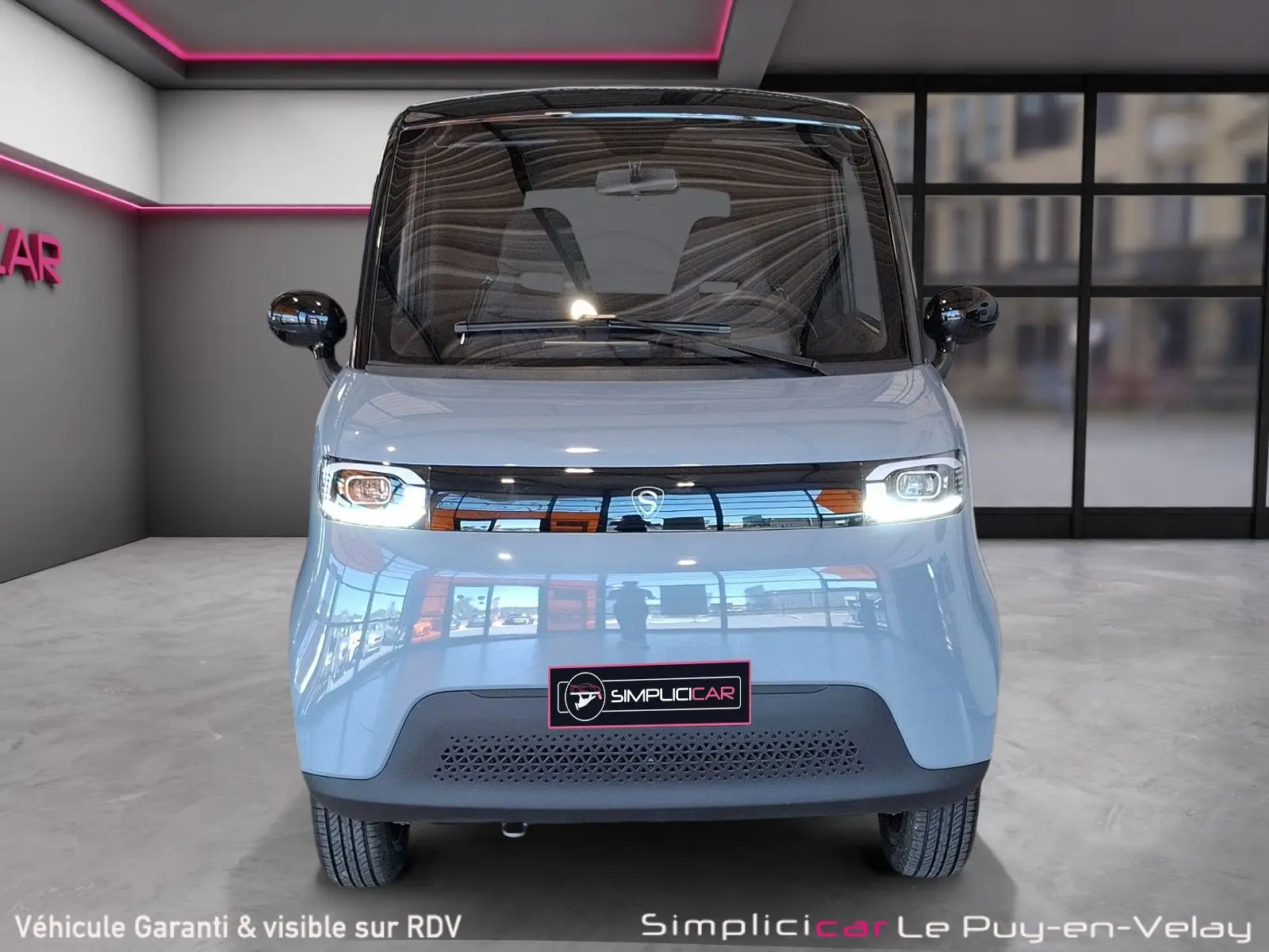 Vue frontale d'une SIMPLICI S1 électrique gris perle avec toit noir et phares allumés dans un showroom moderne.