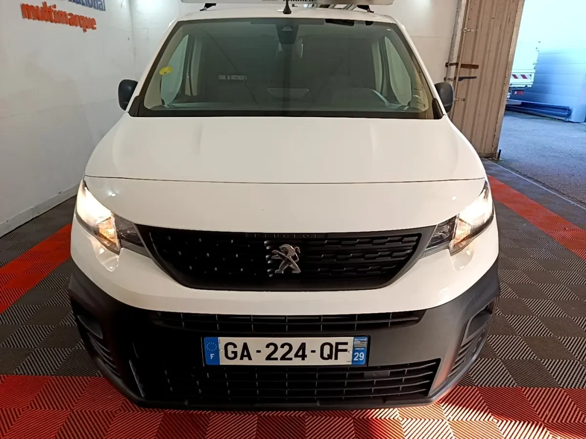 Vue avant d’un Peugeot Partner blanc 2021 avec calandre noire et phares allumés dans un garage.