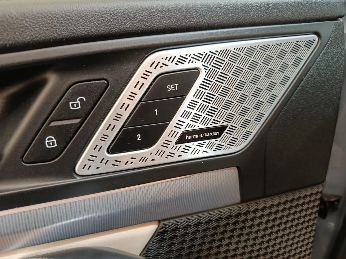 Détail intérieur côté conducteur avec commandes de verrouillage et haut-parleur Harman Kardon argenté sur porte noire.