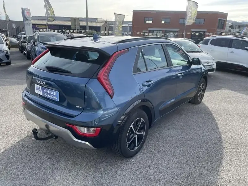 KIA Niro 2024 bleu métal en 3/4 arrière droit, avec feux arrière en forme de C et attelage visible.
