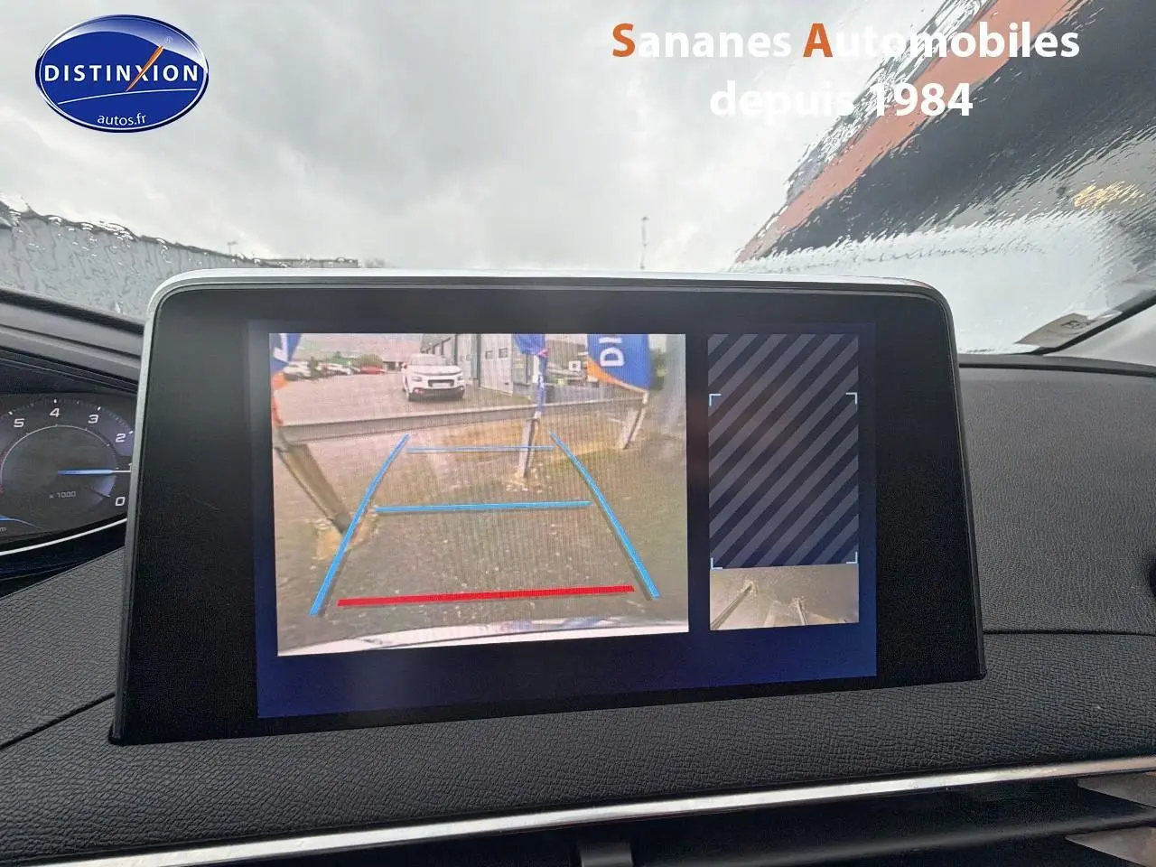 Écran tactile intérieur montrant la caméra de recul d'un Peugeot 3008 blanc, vue frontale sous la pluie.