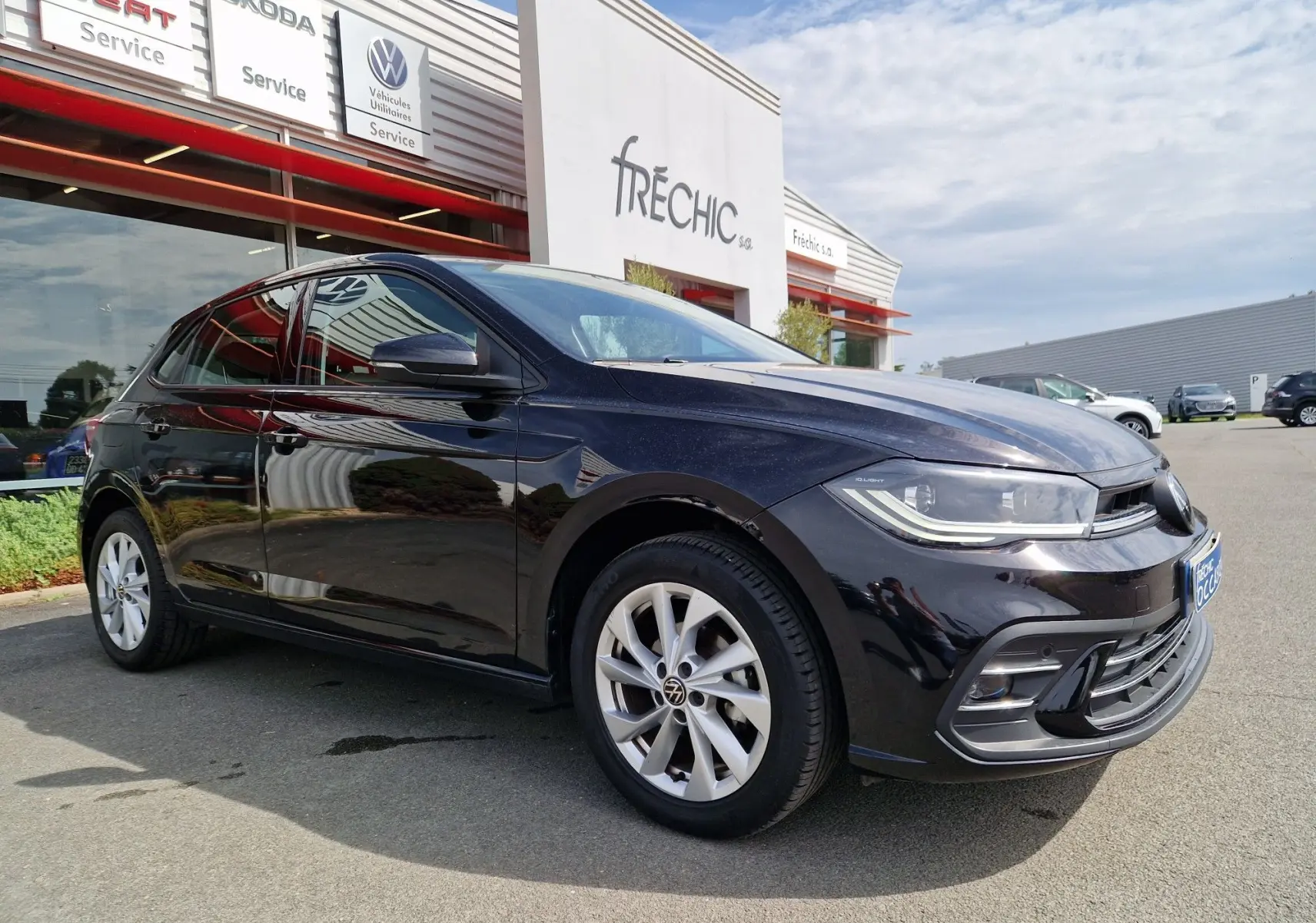 Volkswagen Polo noire vue en 3/4 avant droit, garée devant un concessionnaire avec jantes alliage et phares LED.