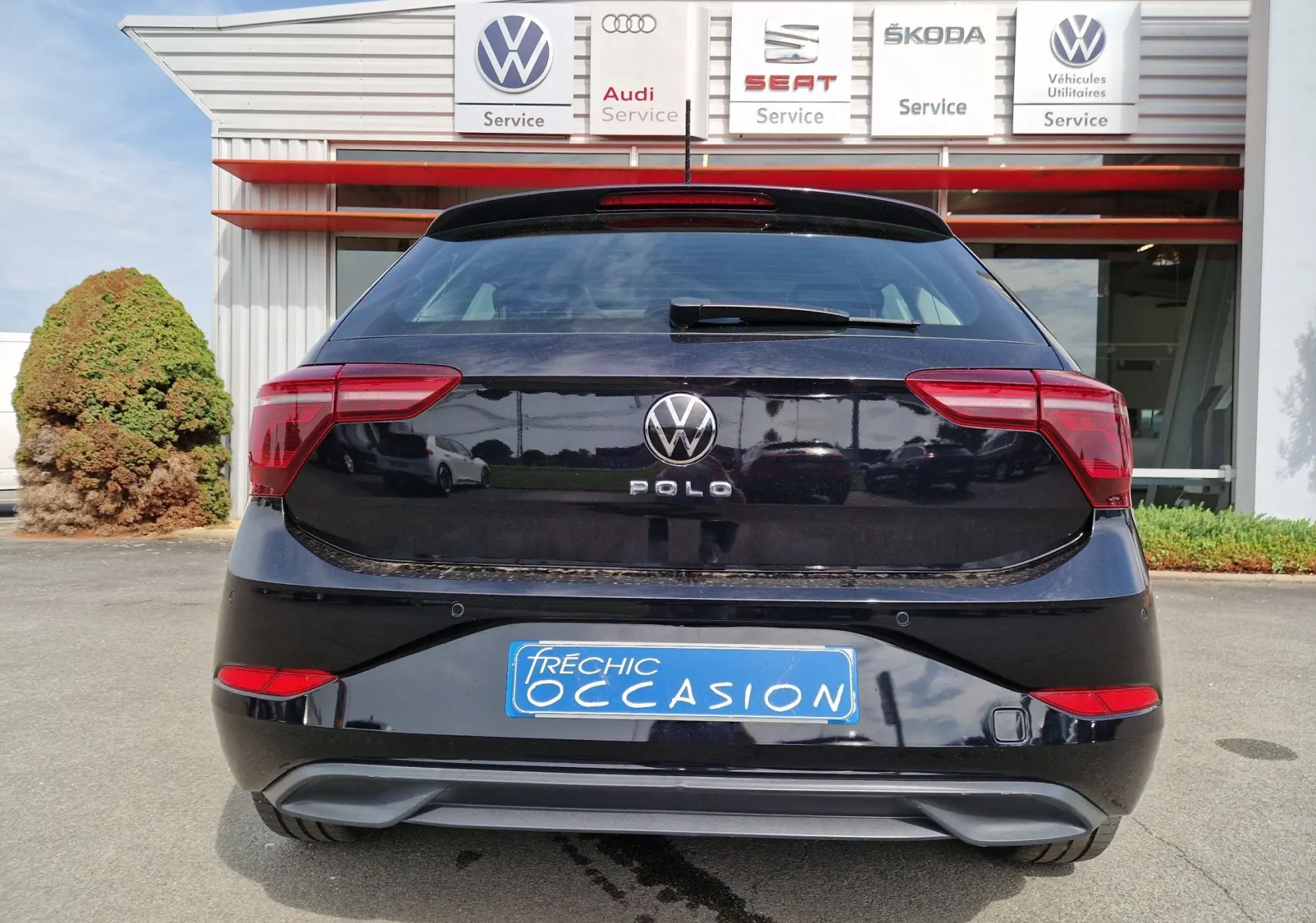 Vue arrière d'une Volkswagen Polo noire 2022 stationnée devant un garage multimarques avec plaque d'occasion bleue.