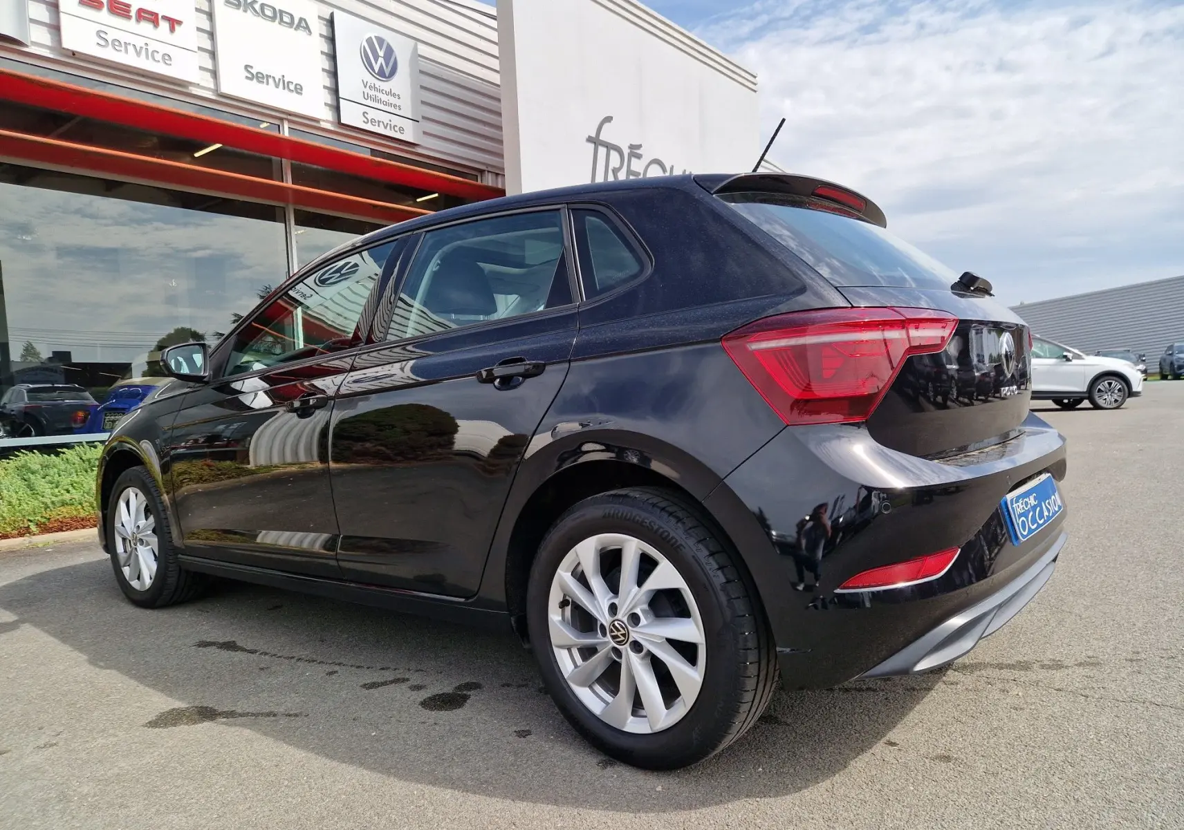 Volkswagen Polo noire vue en 3/4 arrière droit, stationnée devant un service Volkswagen avec jantes alliage.