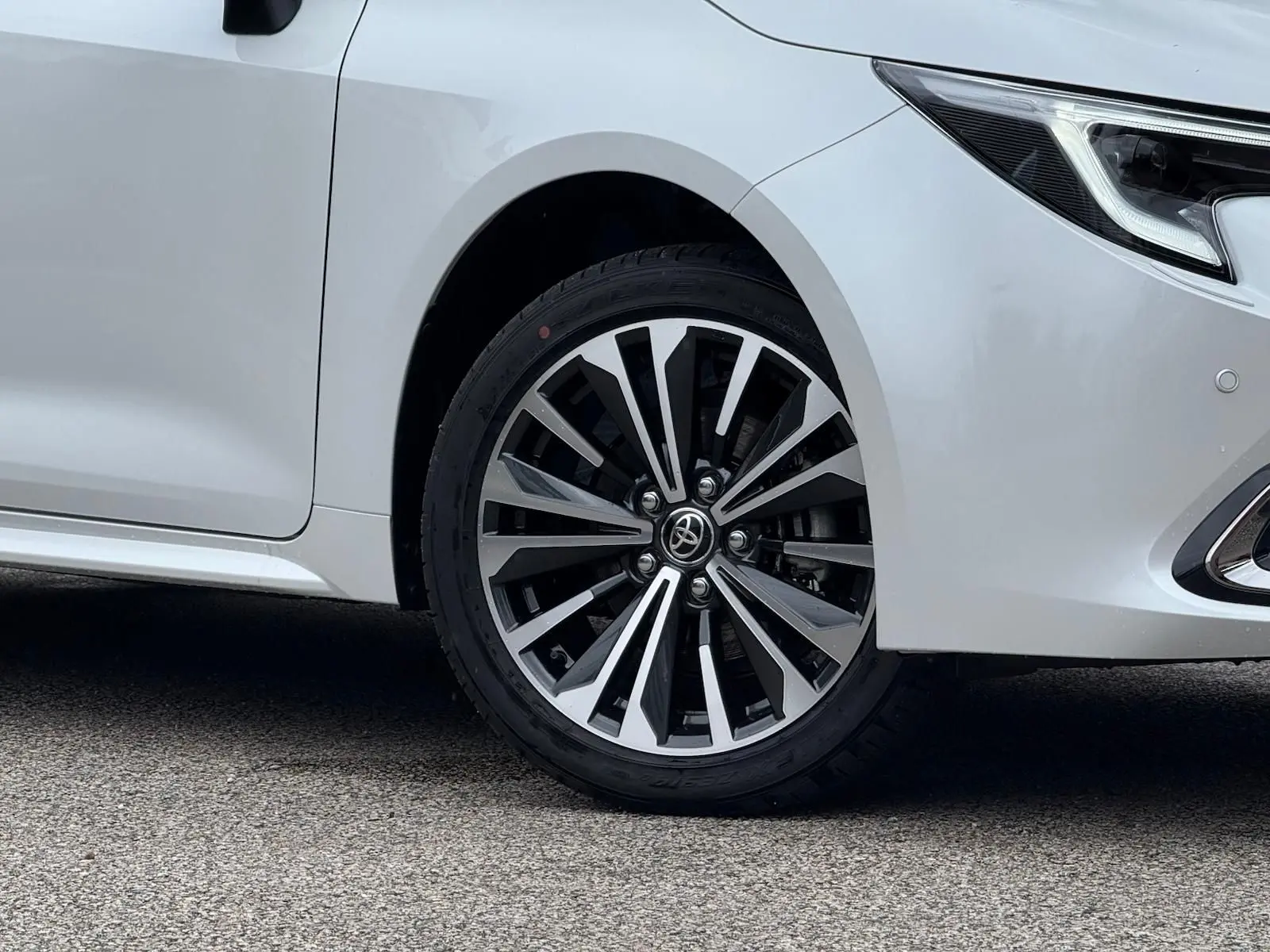 Gros plan sur la roue avant droite et le phare d'une Toyota Corolla Touring Sport blanc lunaire nacré.