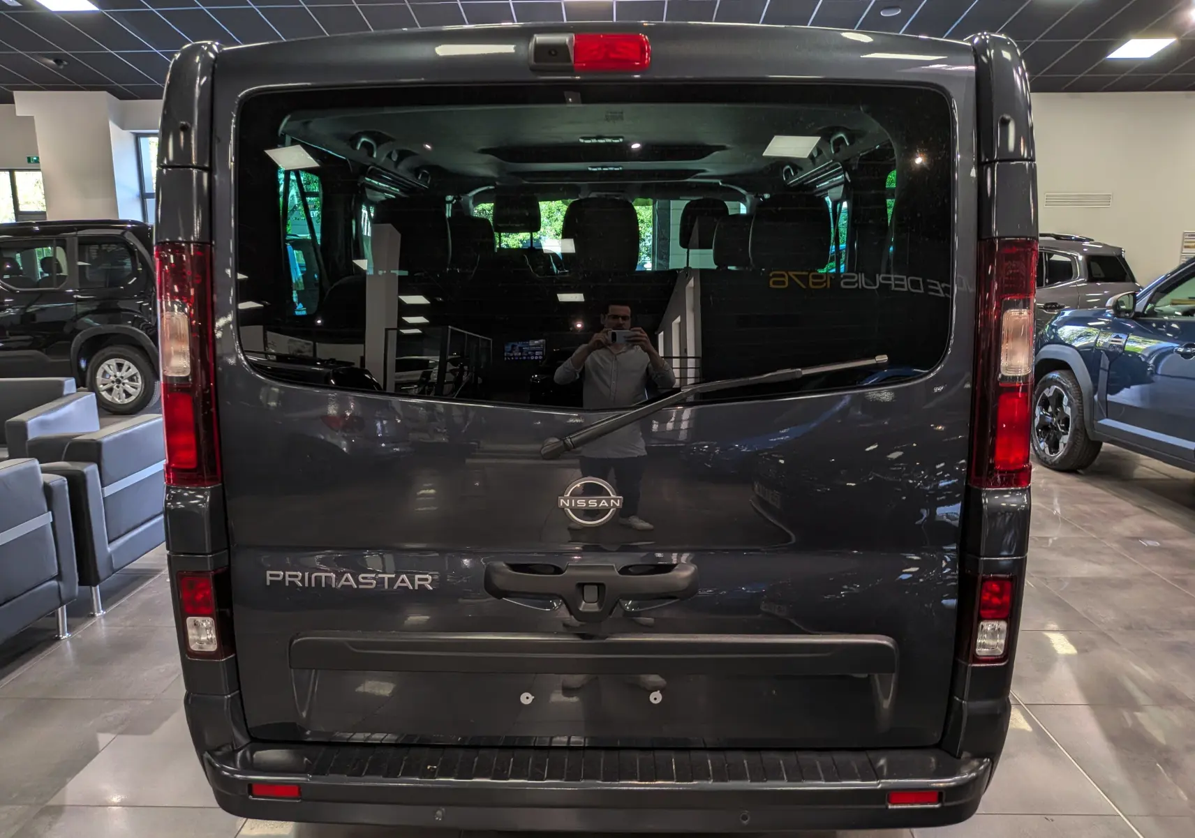 Vue arrière d’un Nissan Primastar Combi gris comète 2025, avec logo et nom du modèle visibles sur la porte arrière vitrée.