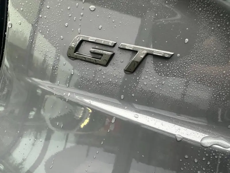 Gros plan sur l'emblème GT argenté sur la carrosserie grise mouillée d'une Peugeot 208 Hybrid 2025.