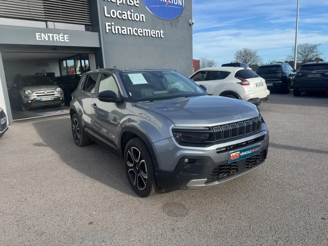 Jeep Avenger gris en 3/4 avant droit, avec jantes alliage noires et calandre distinctive, stationné devant un concessionnaire.
