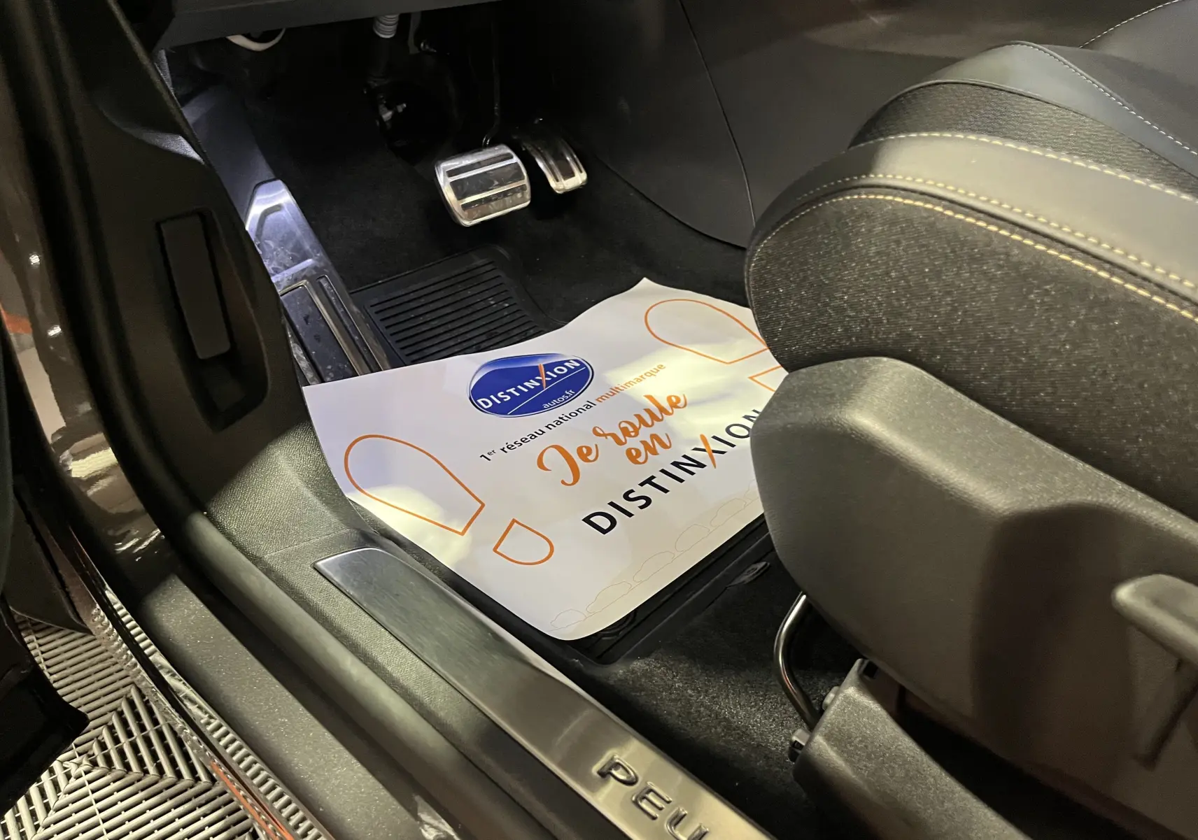 Vue intérieure côté conducteur du Peugeot 5008 gris platinium 2024, avec pédales alu et tapis de sol publicitaire.