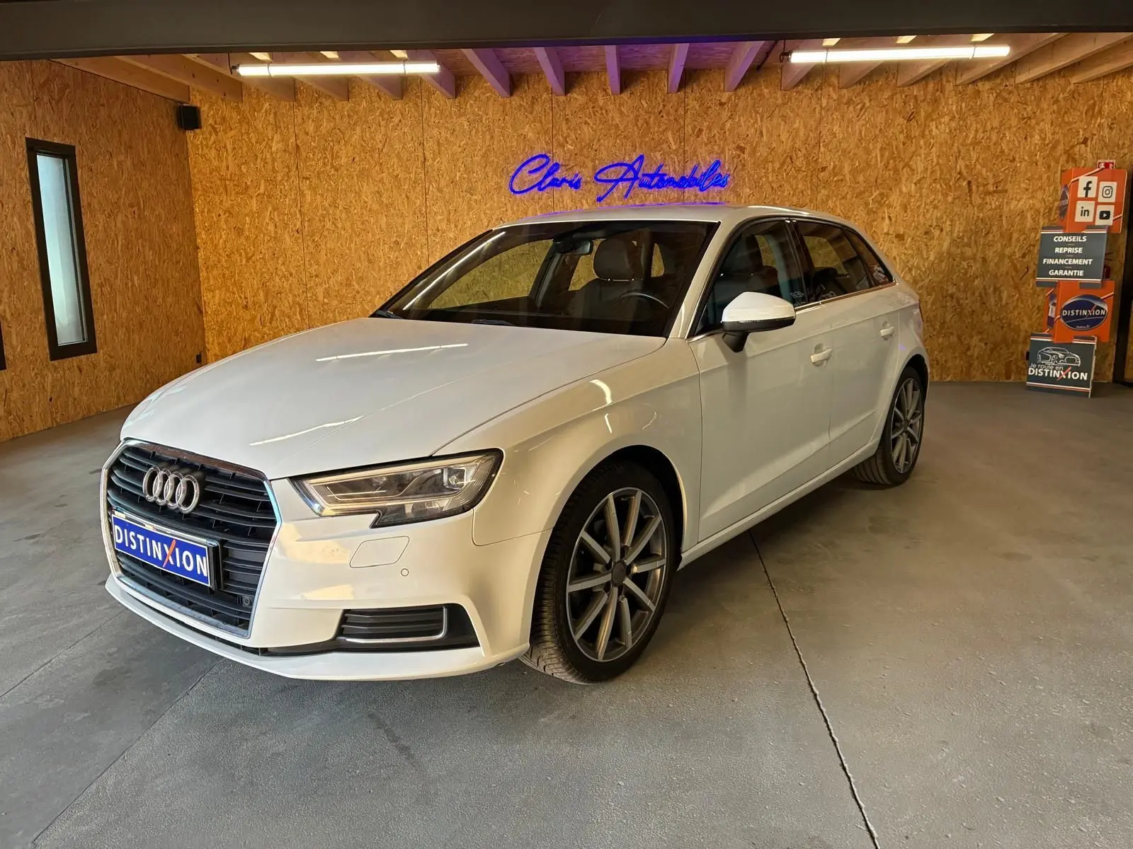 Audi A3 Sportback blanc vue 3/4 avant droit dans un intérieur avec mur en bois et enseigne lumineuse bleue.