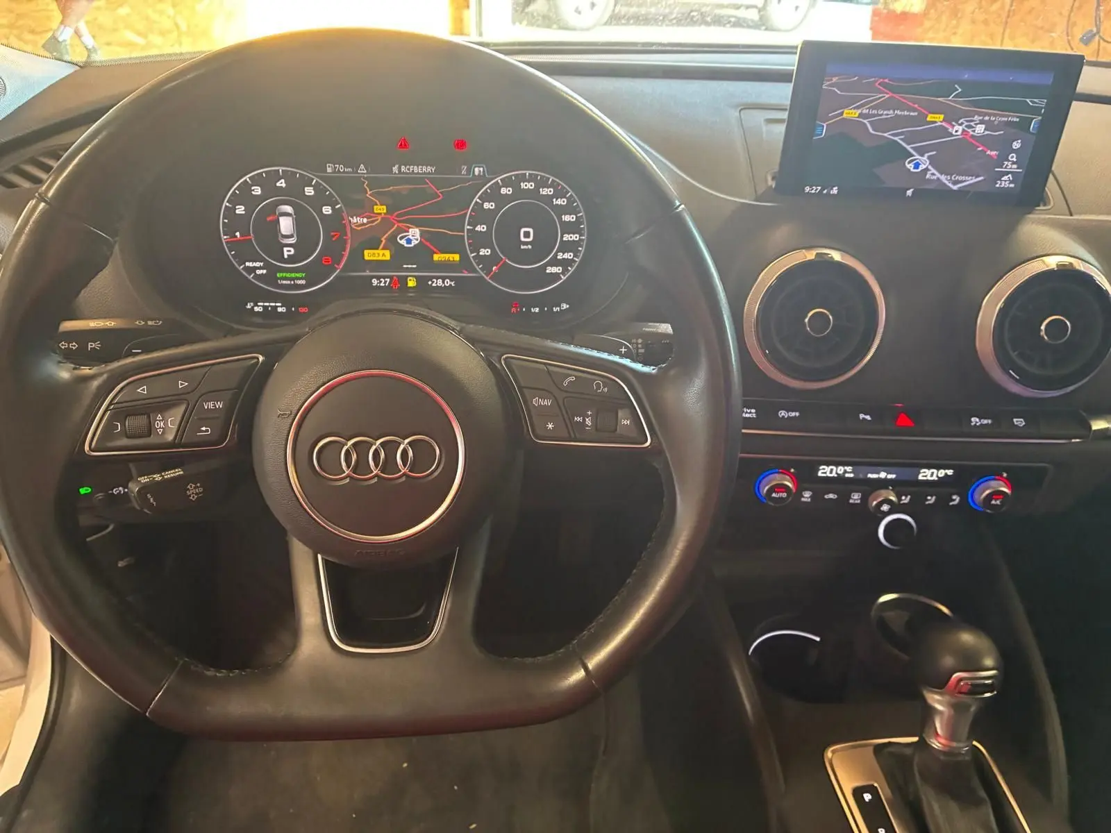 Vue intérieure centrée sur le volant et le tableau de bord noir de l'Audi A3 Sportback avec écran GPS et commandes intégrées.