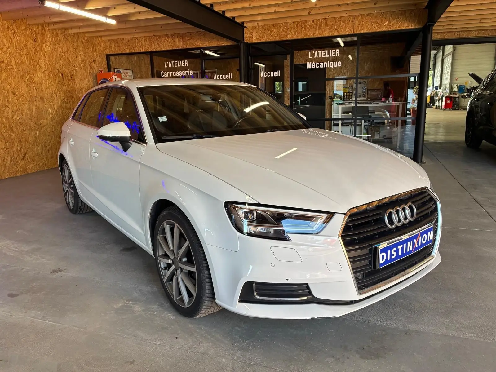 Audi A3 Sportback blanc vue 3/4 avant droit dans un atelier avec calandre noire et jantes alliage.