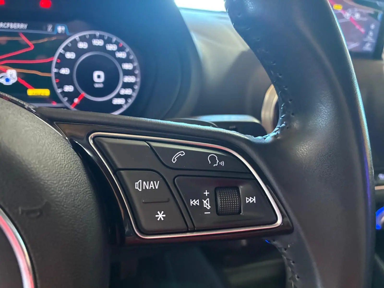 Gros plan sur les commandes audio et navigation au volant en cuir noir de l'Audi A3 Sportback blanc, tableau de bord numérique en arrière-plan.