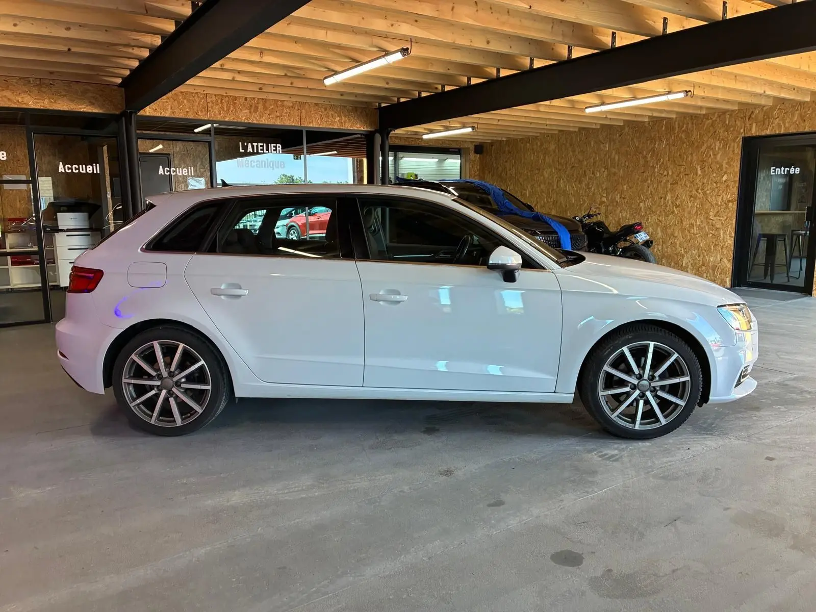 Audi A3 Sportback blanc vue de profil côté gauche, stationnée dans un garage avec intérieur en bois.