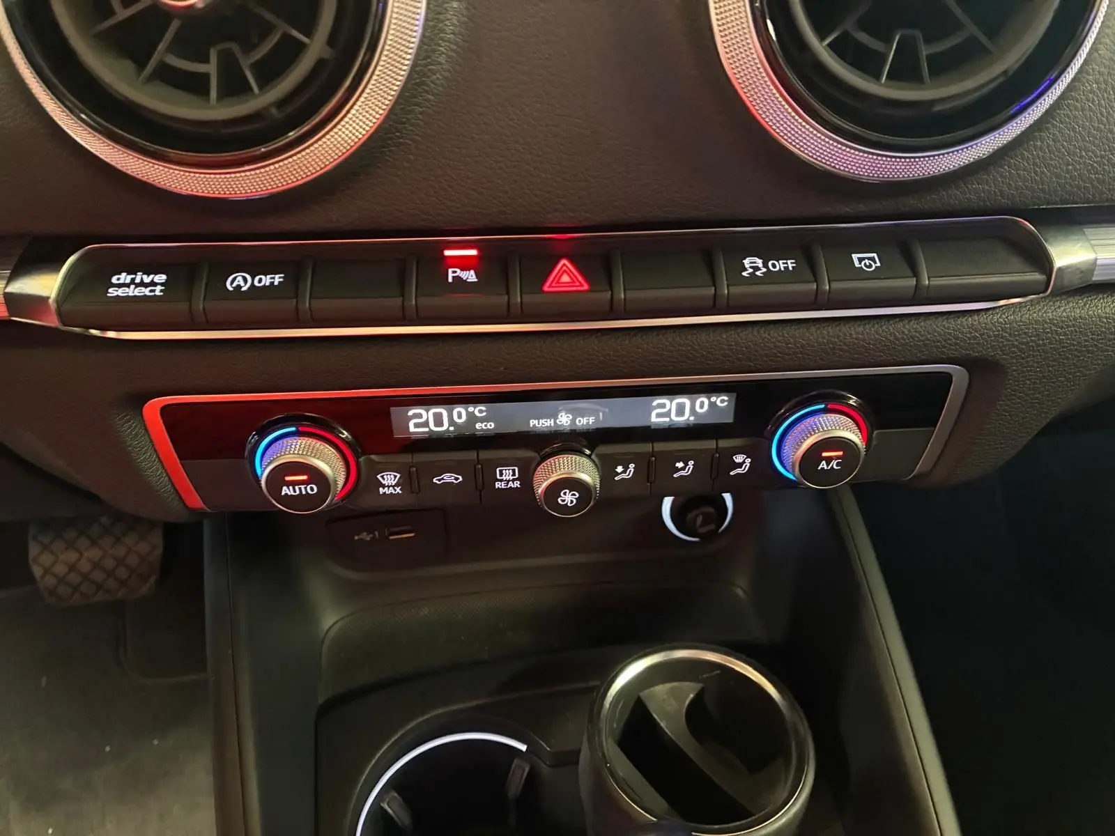 Vue rapprochée du tableau de bord de l’Audi A3 Sportback blanc, avec commandes climatisation et boutons de sécurité éclairés.