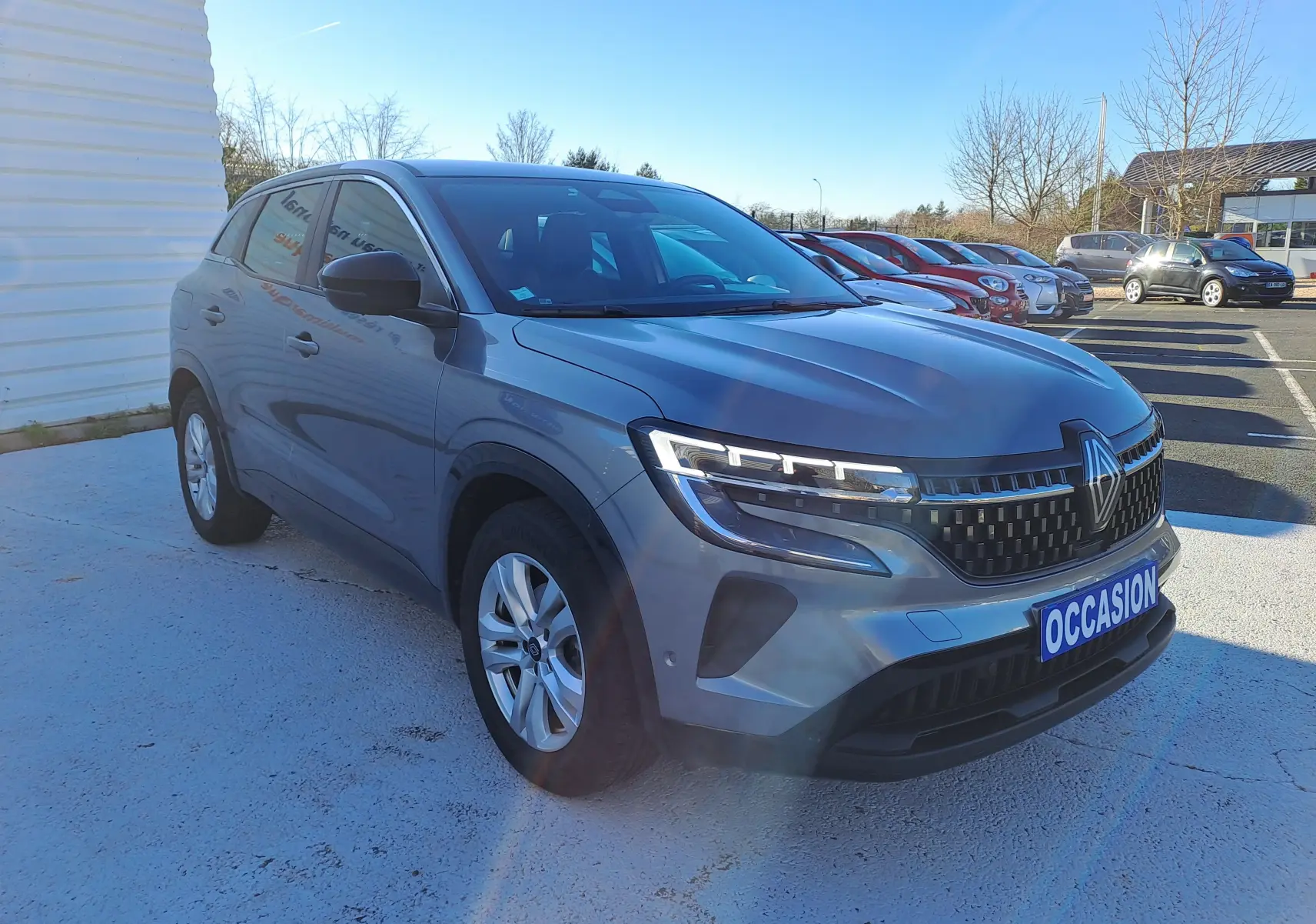 Renault Austral gris schiste en 3/4 avant droit, avec calandre noire et feux LED, sur parking extérieur ensoleillé.