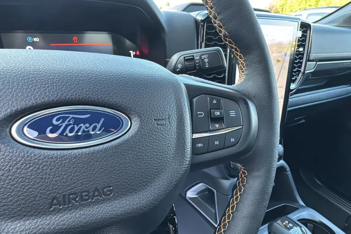 Gros plan sur le volant en cuir noir avec surpiqûres orange du Ford Ranger PHEV 281 Wildtrak, tableau de bord et écran tactile visibles.
