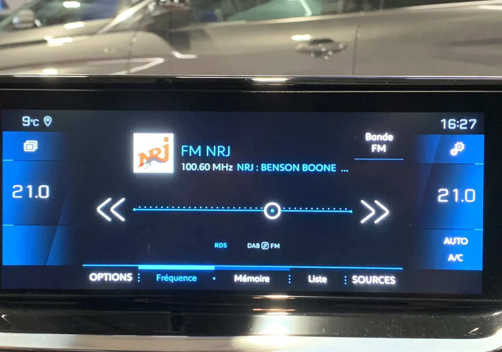 Écran tactile du système multimédia de la Peugeot 208 gris, affichant la radio FM NRJ à 16h27, température 21°C.