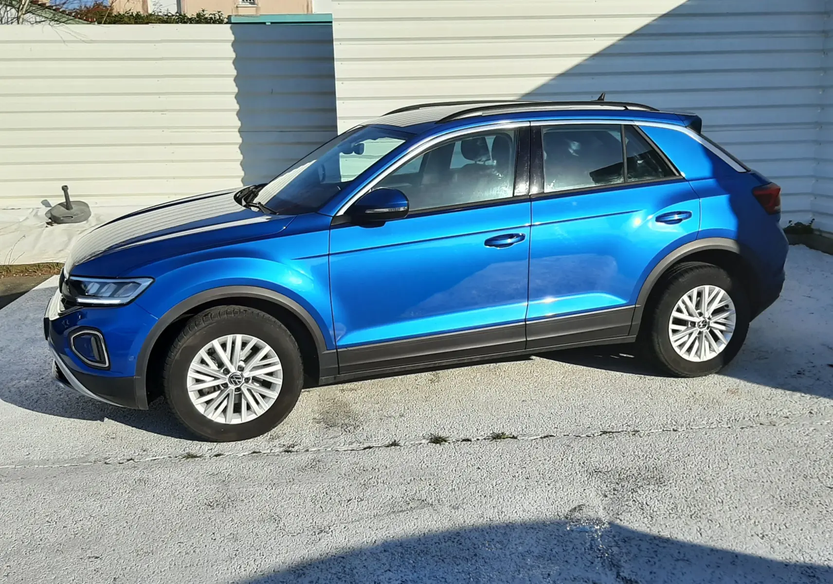Volkswagen T-Roc bleu en profil côté gauche, avec jantes alliage et toit noir sur fond urbain lumineux.