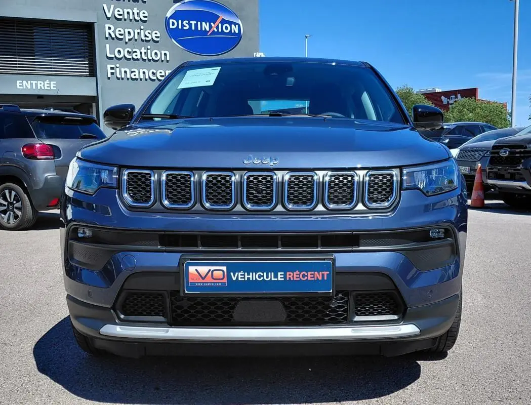 Vue frontale d'un Jeep Compass bleu 2025 avec calandre chromée et plaque "Véhicule récent" sur parking.