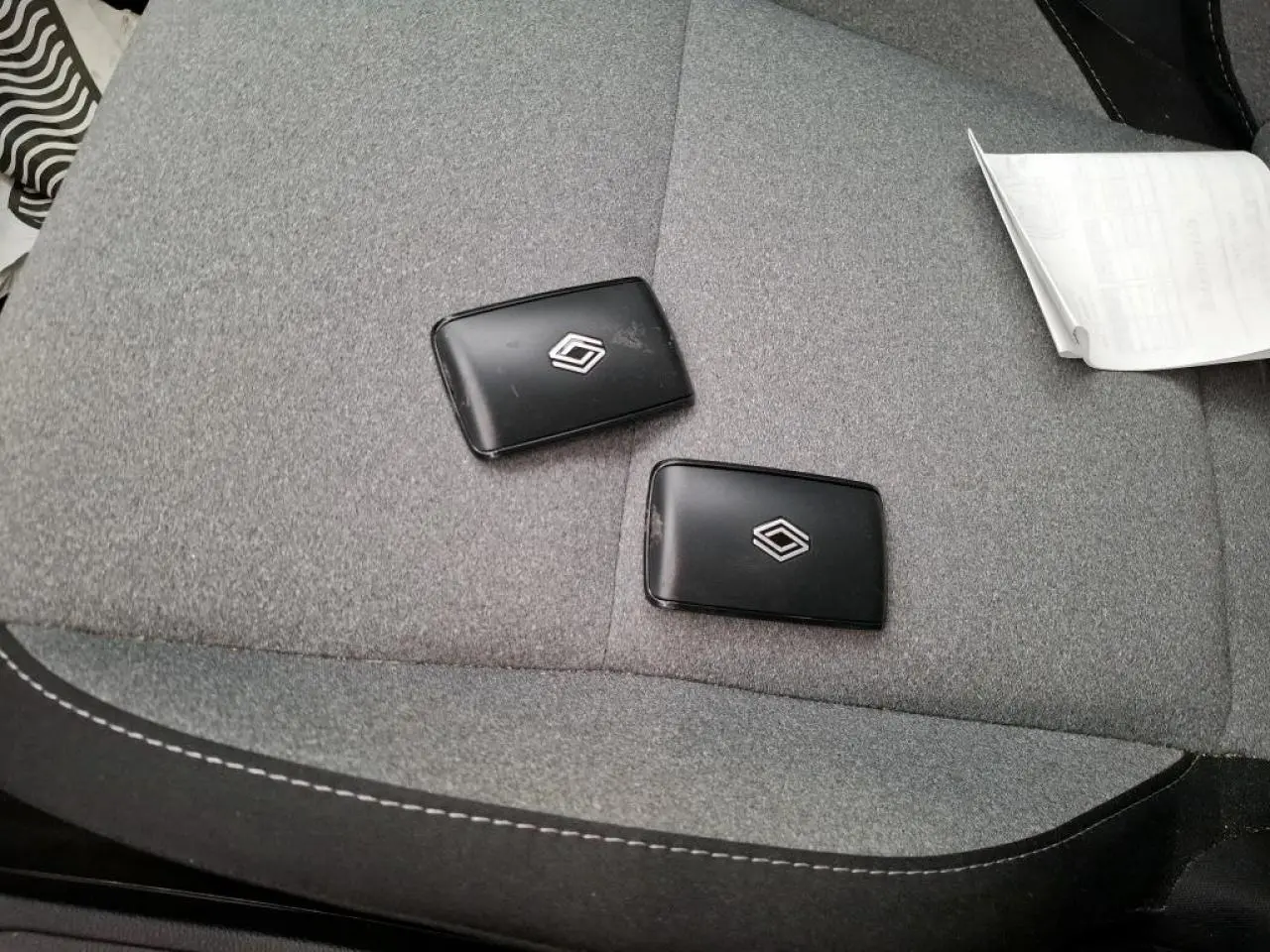 Gros plan sur deux clés noires Renault posées sur un siège tissu gris à coutures blanches, intérieur voiture visible.