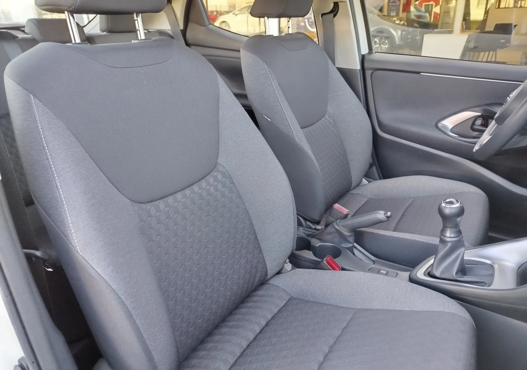 Vue intérieure côté avant droit de la Toyota Yaris blanche, montrant les sièges en tissu gris et le levier de vitesses manuel.