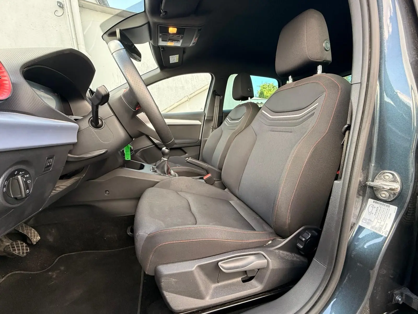 Vue intérieure côté conducteur de la SEAT Ibiza 1.0 TSI 95 FR 2025 en gris foncé, avec sièges tissu noirs et coutures rouges.