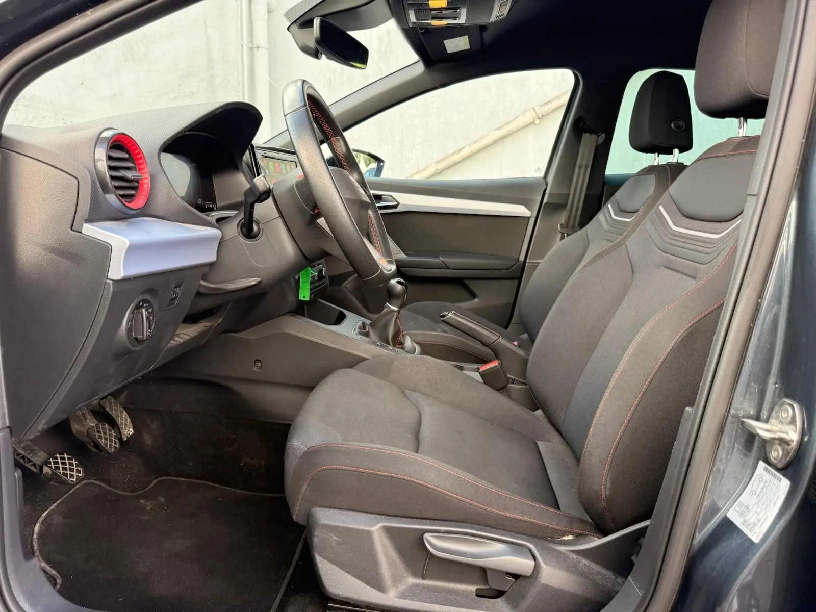 Vue intérieure côté conducteur de la SEAT Ibiza 1.0 TSI 95 FR 2025 avec sièges gris foncé et surpiqûres rouges.