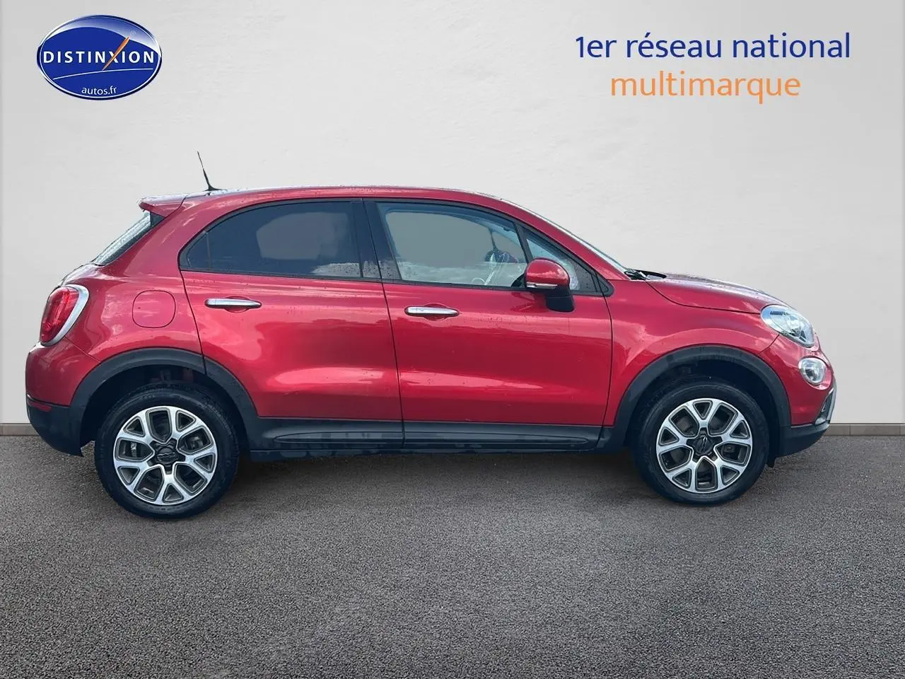 Profil droit d'un FIAT 500X rouge 2016 avec jantes alu et barres de toit, garé devant un mur clair.