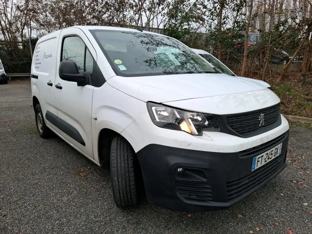 Peugeot Partner blanc vue 3/4 avant gauche avec phare allumé et pare-chocs noir sur parking extérieur.