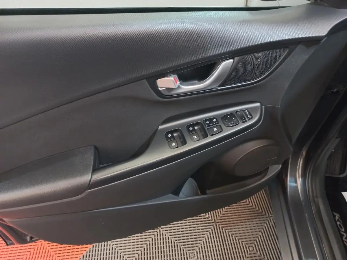 Vue intérieure côté gauche de la porte avant gris foncé du Hyundai Kona 1.6 Hybrid avec commandes électriques et poignée métal.