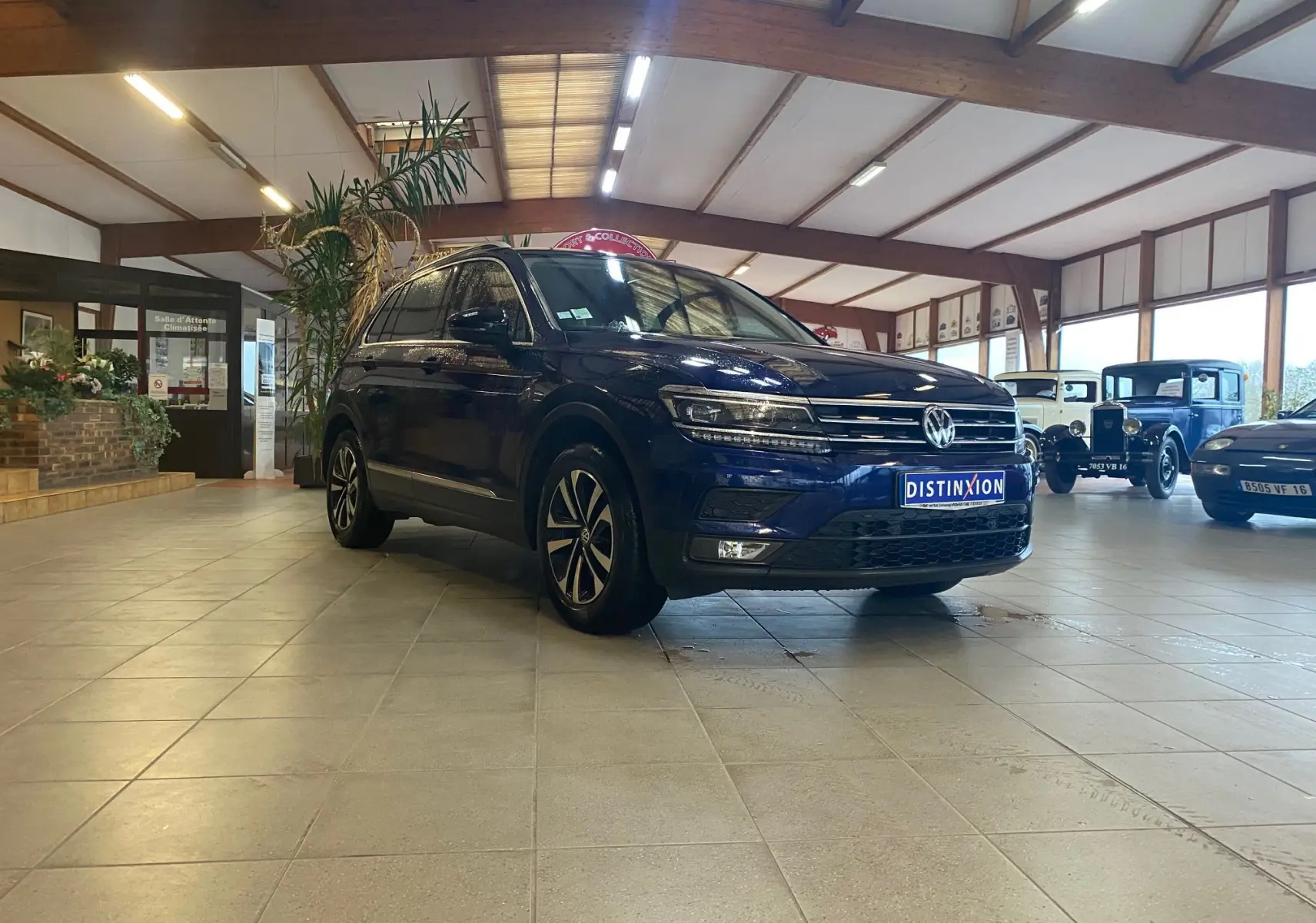 Volkswagen Tiguan bleu vert 2020 vu en 3/4 avant droit dans un showroom avec voitures anciennes en arrière-plan.