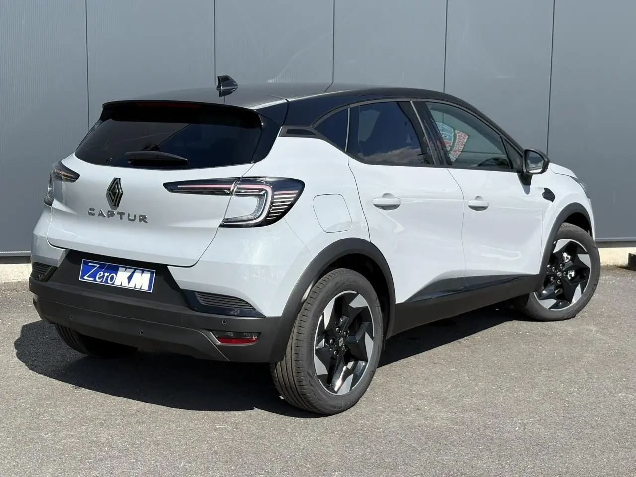 Vue 3/4 arrière droit d'un Renault Captur blanc avec toit noir et jantes noires sur fond gris urbain.