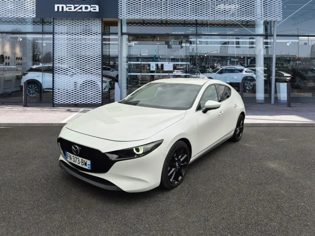 Vue 3/4 avant d'une Mazda3 blanche 2020 avec jantes noires, stationnée devant une concession Mazda.