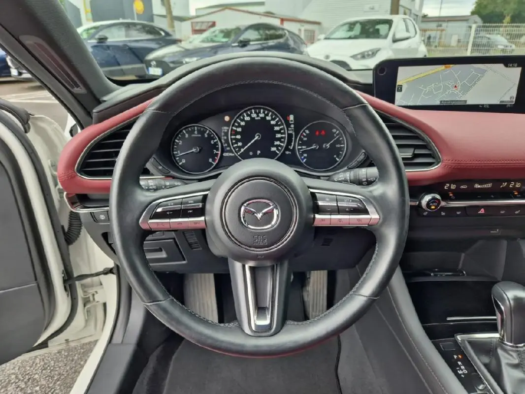 Vue intérieure centrée sur le volant noir d'une Mazda3 2020 avec tableau de bord rouge et écran GPS intégré.