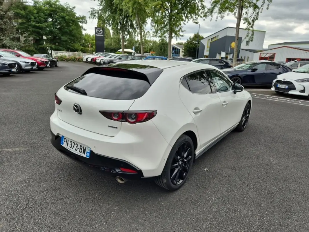 Vue 3/4 arrière droite d'une Mazda3 blanche 2020 avec jantes noires et vitres teintées dans un parking extérieur.