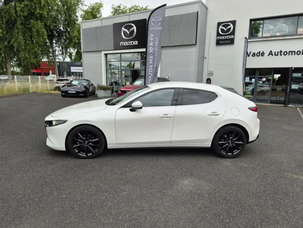 Vue de profil côté gauche d'une Mazda3 blanche avec jantes noires, stationnée devant un concessionnaire Mazda.