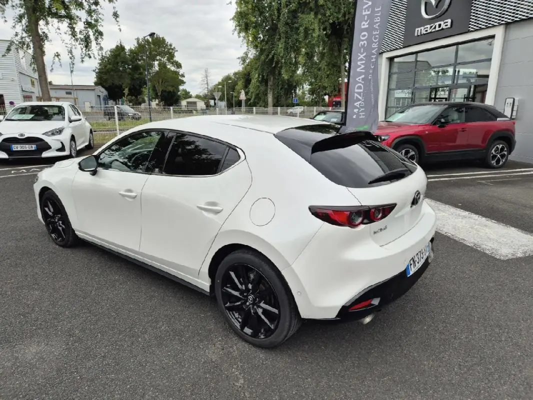 Vue 3/4 arrière droite d'une Mazda3 blanche 2020 avec jantes noires et vitres teintées, stationnée devant un concessionnaire Mazda.