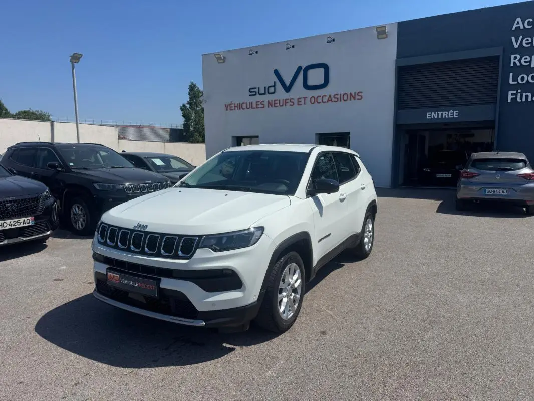 Jeep Compass blanc vue 3/4 avant droit, stationné devant un bâtiment de vente de véhicules.